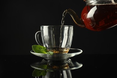 Photo of Pouring freshly brewed tea from teapot into cup at dark mirror table, closeup
