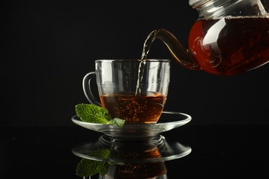 Photo of Pouring freshly brewed tea from teapot into cup at dark mirror table, closeup