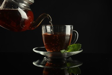 Photo of Pouring freshly brewed tea from teapot into cup at dark mirror table, closeup