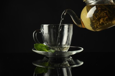 Photo of Pouring freshly brewed tea from teapot into cup at dark mirror table, closeup