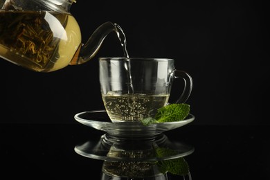 Photo of Pouring freshly brewed tea from teapot into cup at dark mirror table, closeup