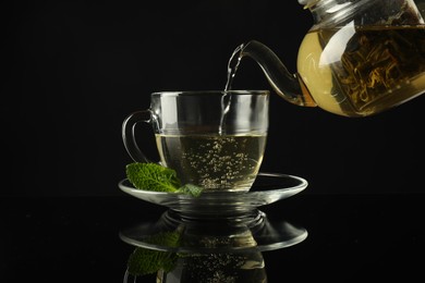 Photo of Pouring freshly brewed tea from teapot into cup at dark mirror table, closeup