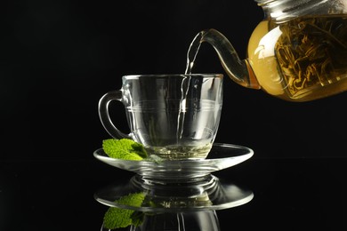 Photo of Pouring freshly brewed tea from teapot into cup at dark mirror table, closeup