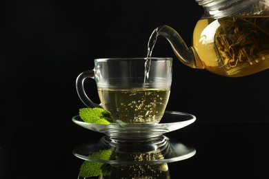 Photo of Pouring freshly brewed tea from teapot into cup at dark mirror table, closeup