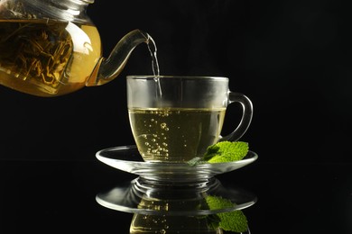Photo of Pouring freshly brewed tea from teapot into cup at dark mirror table, closeup