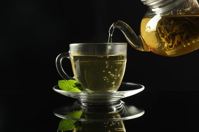 Photo of Pouring freshly brewed tea from teapot into cup at dark mirror table, closeup