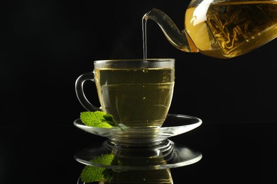 Photo of Pouring freshly brewed tea from teapot into cup at dark mirror table, closeup
