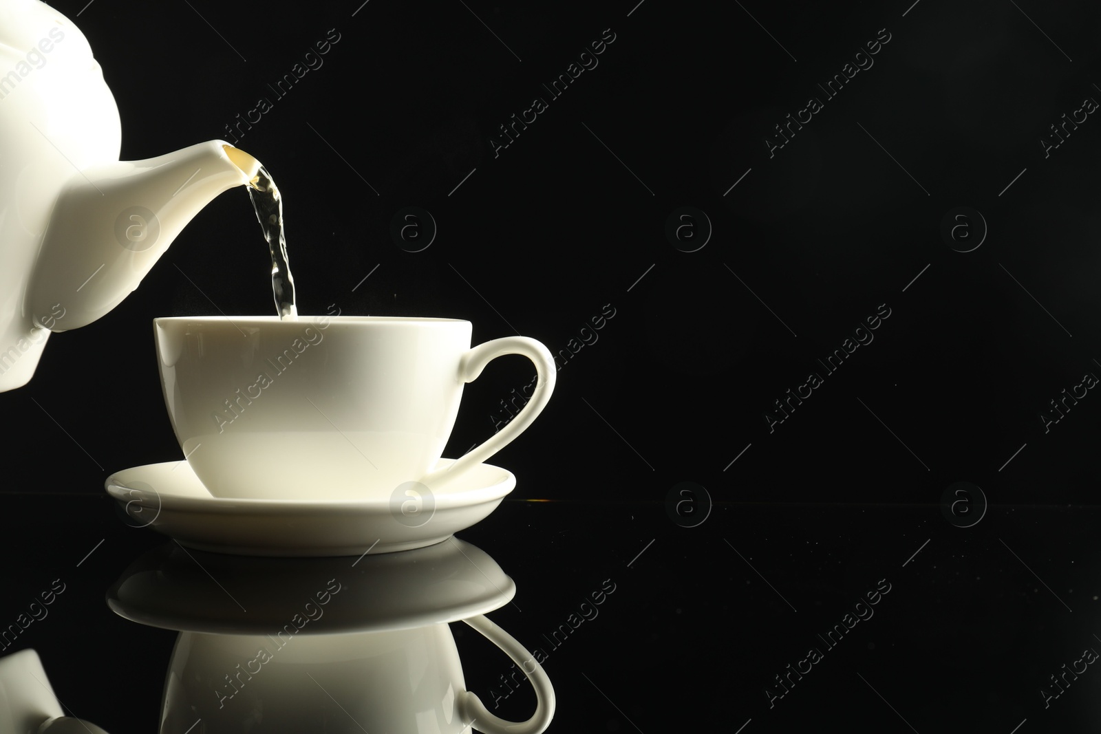 Pouring freshly brewed tea from teapot into cup at dark mirror table, closeup. Space for text Photo of Pouring freshly brewed tea from teapot into cup at dark mirror table, closeup. Space for text