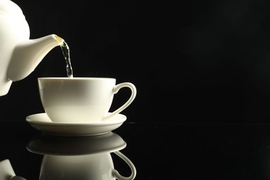 Photo of Pouring freshly brewed tea from teapot into cup at dark mirror table, closeup. Space for text