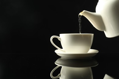 Photo of Pouring freshly brewed tea from teapot into cup at dark mirror table, closeup. Space for text