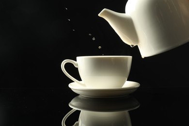 Photo of Pouring freshly brewed tea from teapot into cup at dark mirror table, closeup