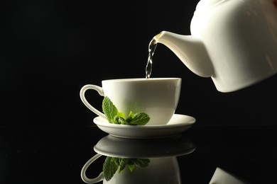 Photo of Pouring freshly brewed tea from teapot into cup at dark mirror table, closeup