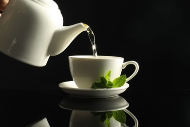 Photo of Pouring freshly brewed tea from teapot into cup at dark mirror table, closeup