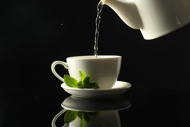 Photo of Pouring freshly brewed tea from teapot into cup at dark mirror table, closeup