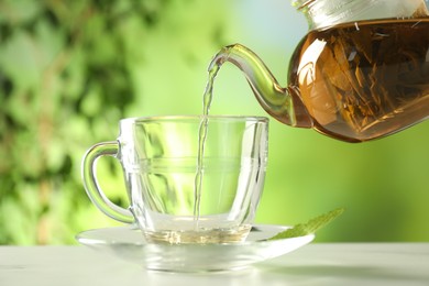 Photo of Pouring freshly brewed tea from teapot into cup at white marble table outdoors, closeup