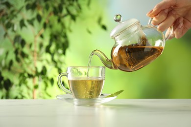 Photo of Woman pouring freshly brewed tea from teapot into cup at white marble table outdoors, closeup
