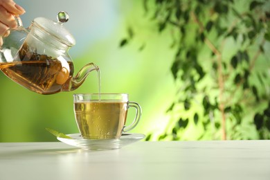 Photo of Woman pouring freshly brewed tea from teapot into cup at white marble table outdoors, closeup. Space for text