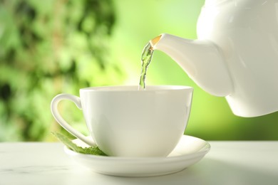 Photo of Pouring freshly brewed tea from teapot into cup at white marble table outdoors, closeup