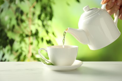 Photo of Woman pouring freshly brewed tea from teapot into cup at white marble table outdoors, closeup
