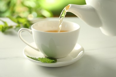 Photo of Pouring freshly brewed tea from teapot into cup at white marble table, closeup