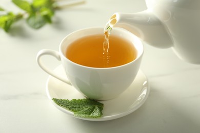 Photo of Pouring freshly brewed tea from teapot into cup at white marble table, closeup