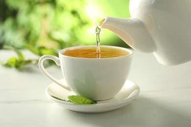Photo of Pouring freshly brewed tea from teapot into cup at white marble table, closeup