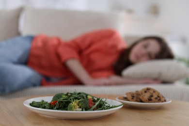 Photo of Sad teen girl lying on sofa near table with salad and cookies at home, selective focus. Eating disorder