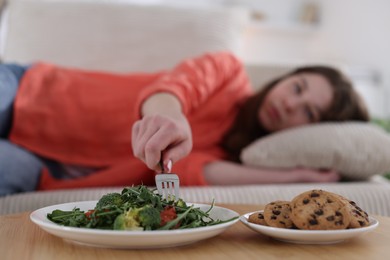 Photo of Sad teen girl taking broccoli from plate while lying on sofa at home, selective focus. Eating disorder