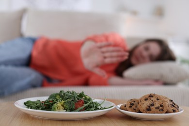 Photo of Sad teen girl showing stop gesture near table with salad and cookies at home, selective focus. Eating disorder