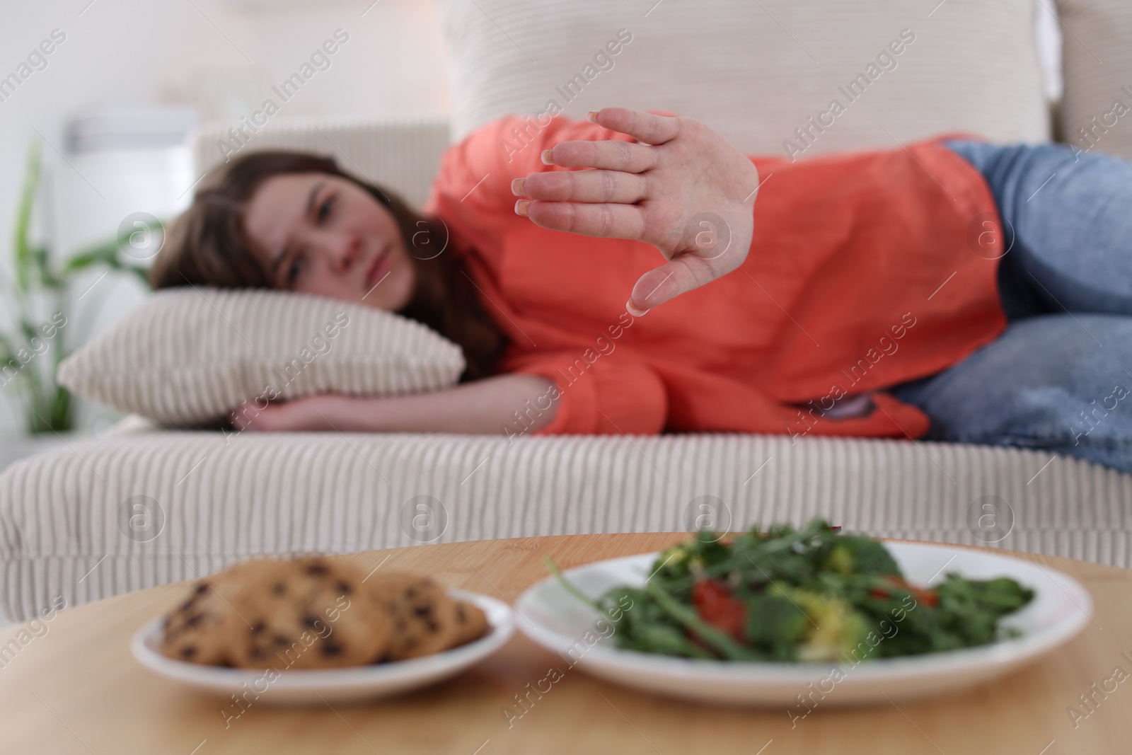 Photo of Sad teen girl showing stop gesture near table with salad and cookies at home, selective focus. Eating disorder