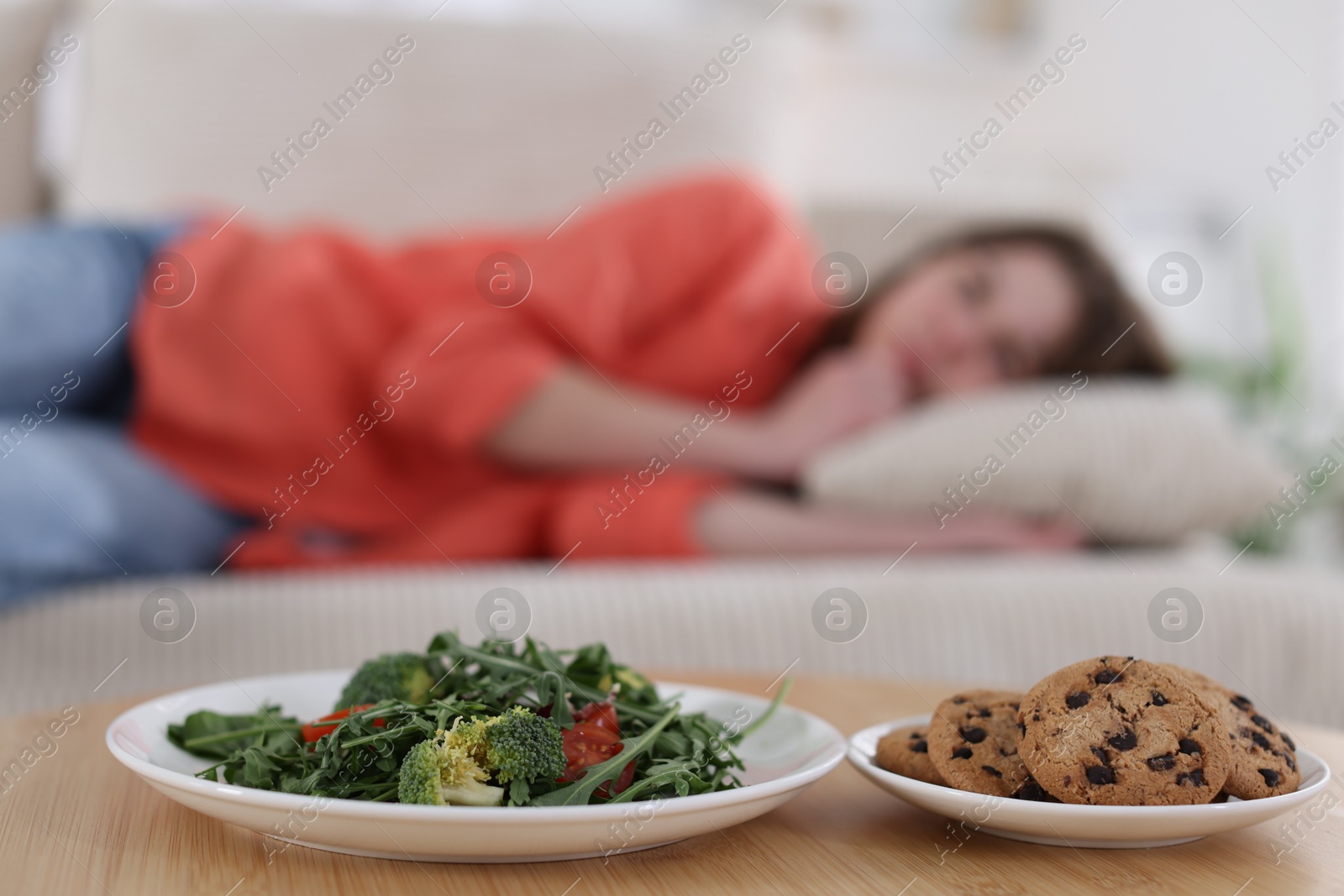 Photo of Sad teen girl lying on sofa near table with salad and cookies at home, selective focus. Eating disorder