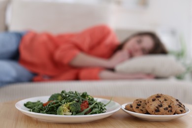 Photo of Sad teen girl lying on sofa near table with salad and cookies at home, selective focus. Eating disorder