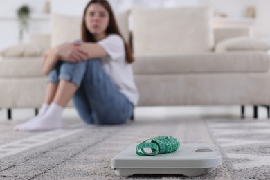 Photo of Sad teen girl sitting on rug near scales and measuring tape at home, selective focus. Eating disorder