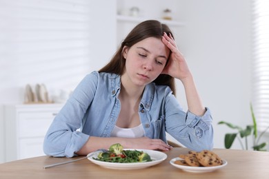 Photo of Sad teen girl at table with salad and cookies indoors. Eating disorder