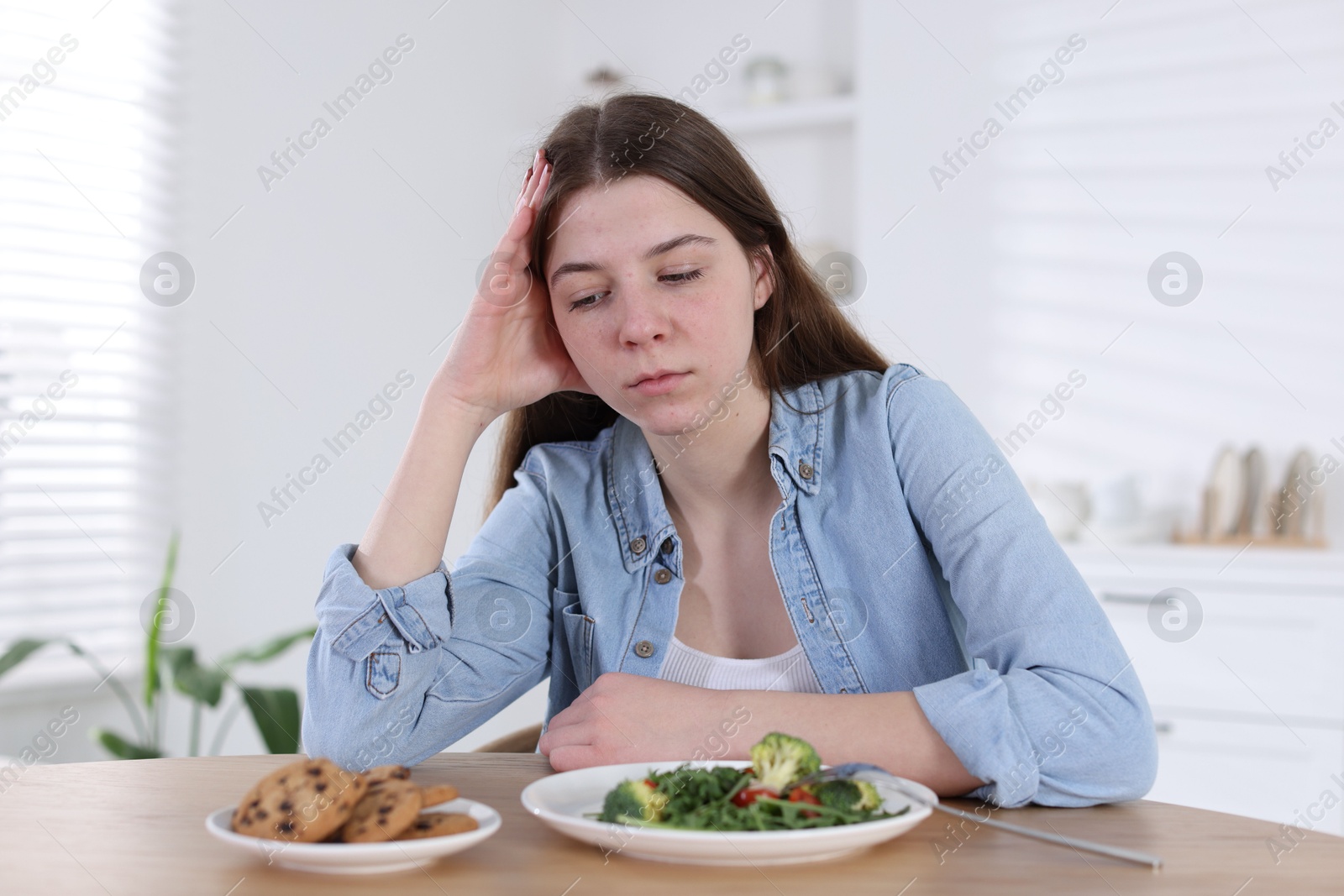 Photo of Sad teen girl at table with salad and cookies indoors. Eating disorder