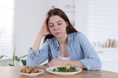 Photo of Sad teen girl at table with salad and cookies indoors. Eating disorder