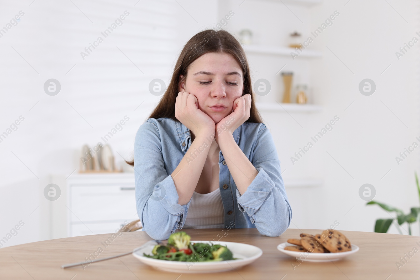 Photo of Sad teen girl at table with salad and cookies indoors. Eating disorder