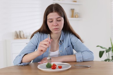 Photo of Sad teen girl holding fork with broccoli at home. Eating disorder