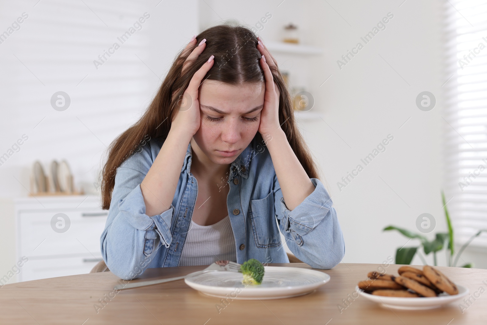 Photo of Sad teen girl at table with broccoli indoors. Eating disorder