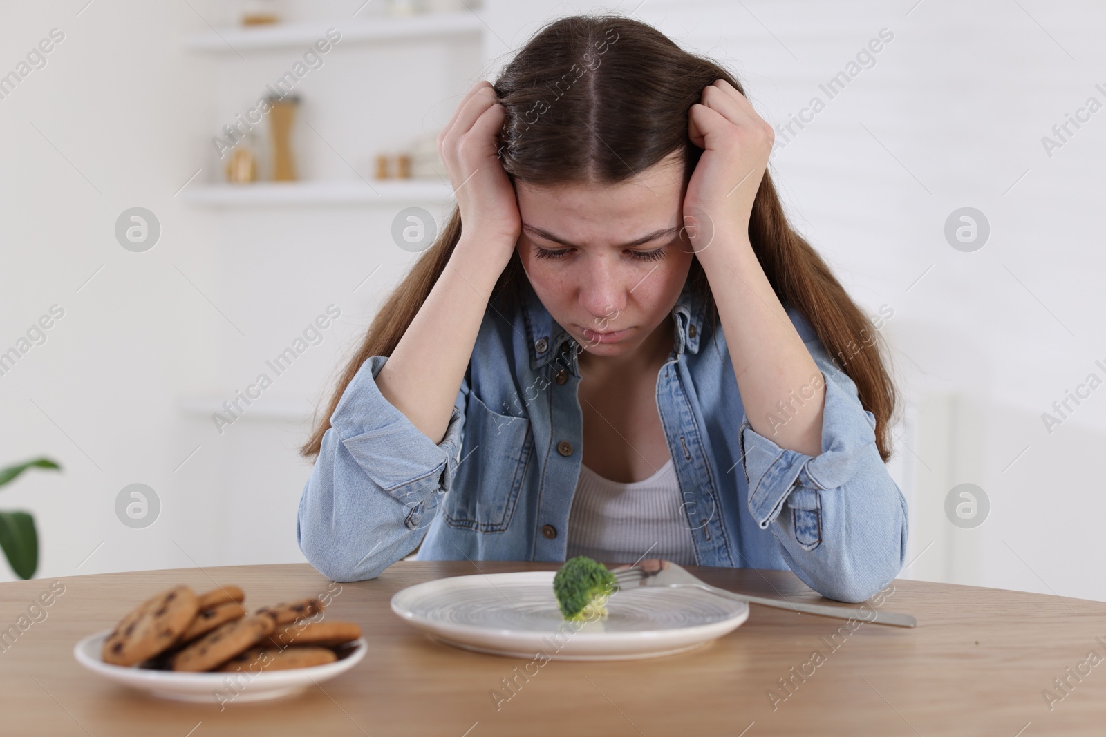 Photo of Sad teen girl at table with broccoli indoors. Eating disorder