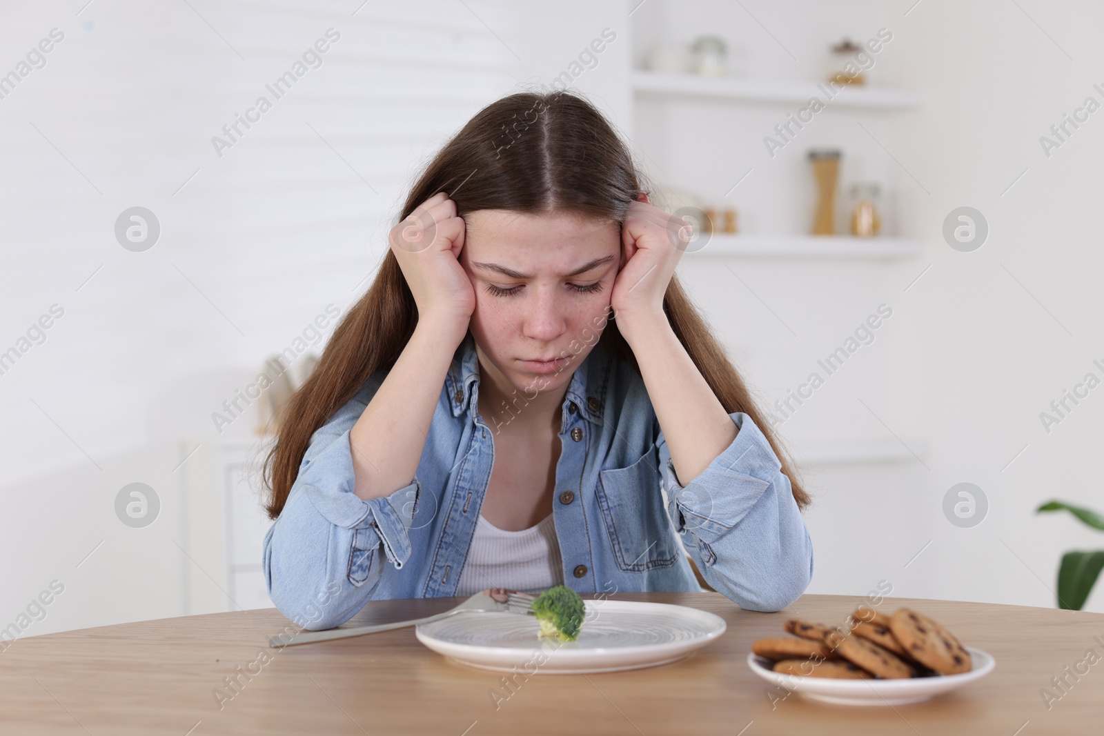 Photo of Sad teen girl at table with broccoli indoors. Eating disorder