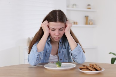 Photo of Sad teen girl at table with broccoli indoors. Eating disorder