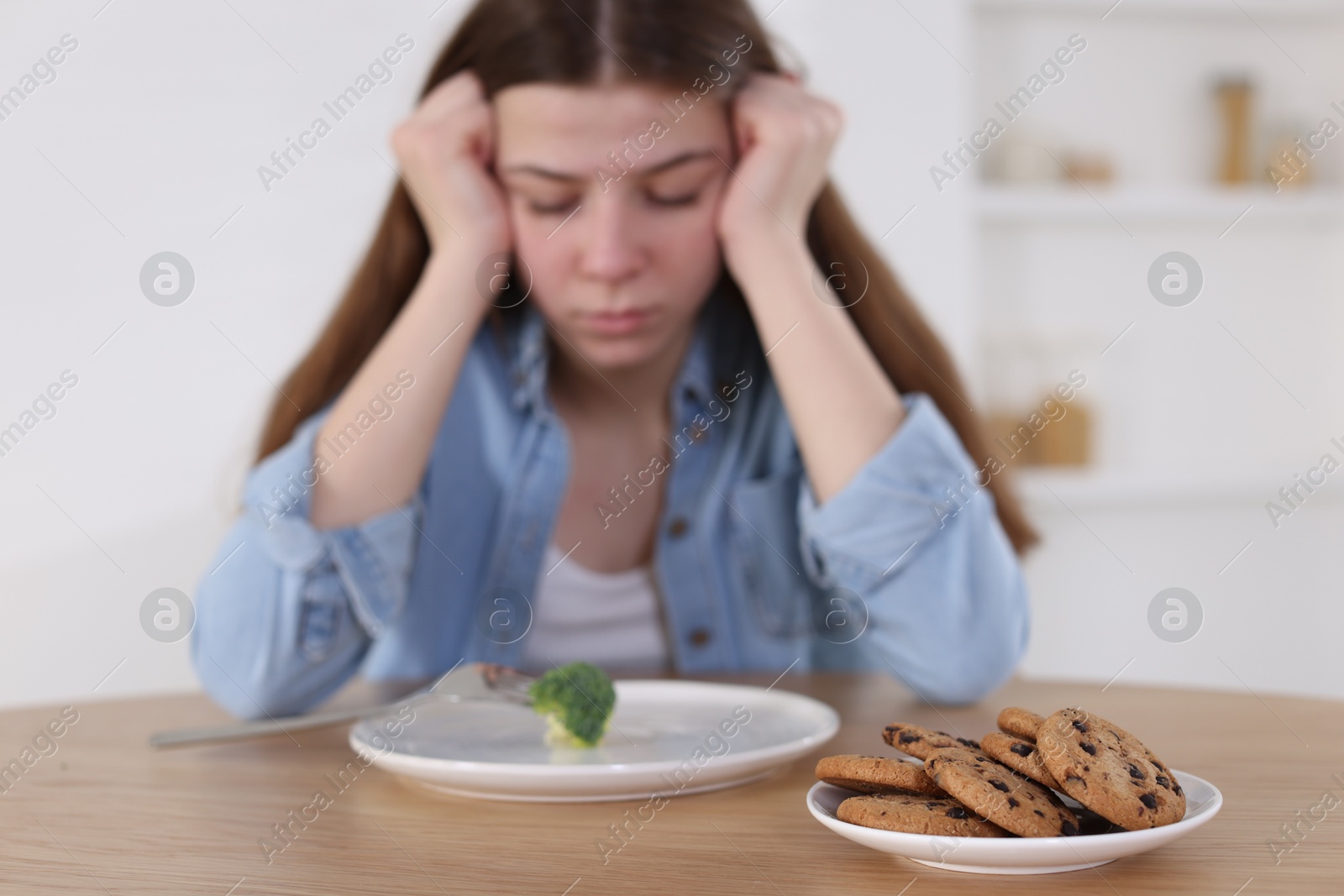 Photo of Sad teen girl at table with broccoli indoors, focus on cookies. Eating disorder