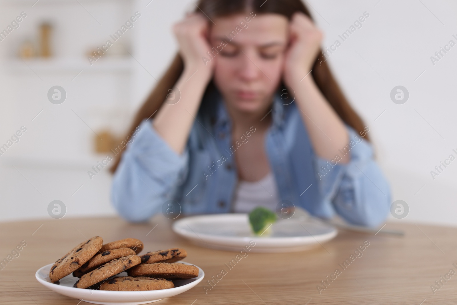 Photo of Sad teen girl at table with broccoli indoors, focus on cookies. Eating disorder