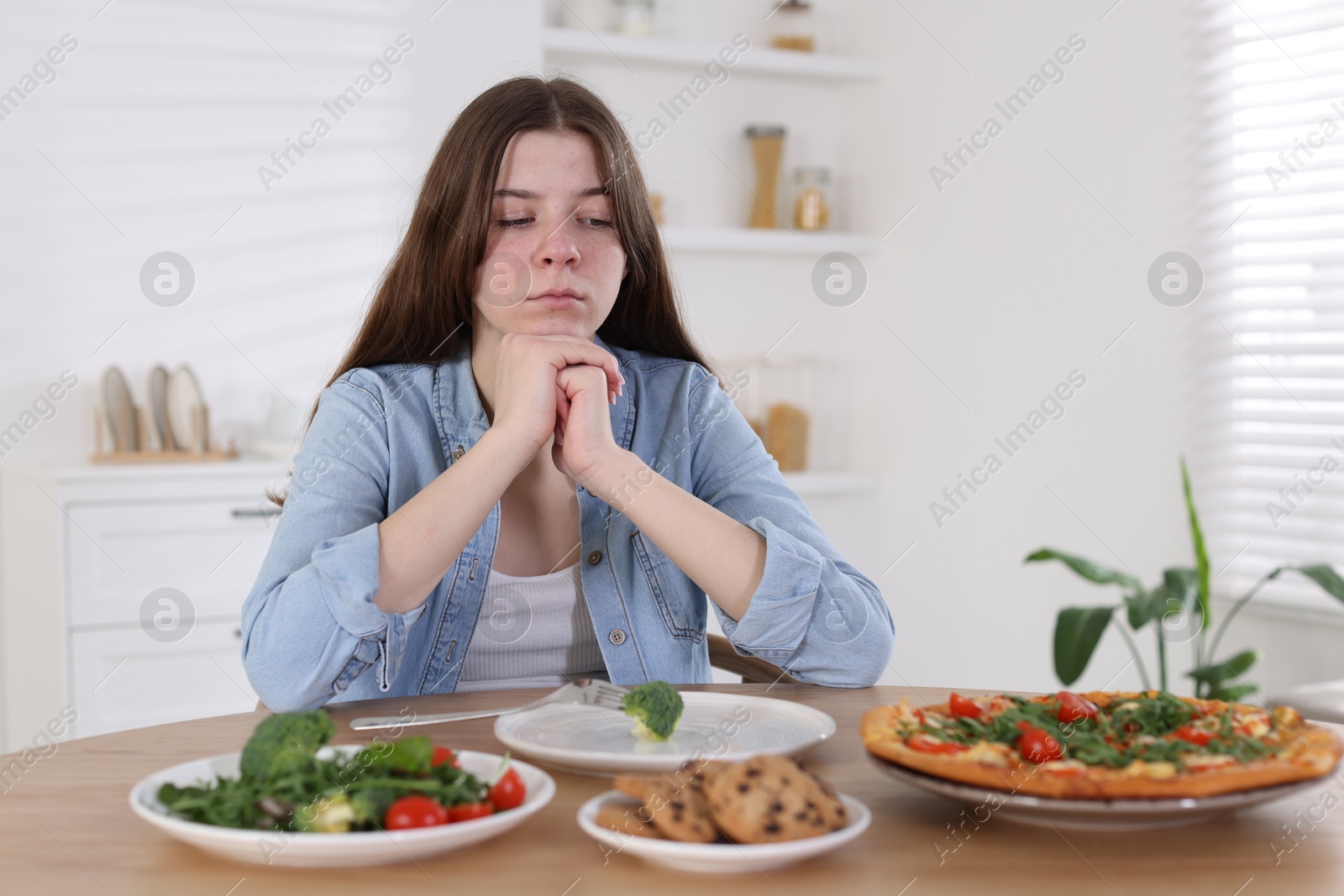 Photo of Sad teen girl at table with pizza, salad and cookies indoors. Eating disorder