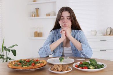 Photo of Sad teen girl at table with pizza, salad and cookies indoors. Eating disorder