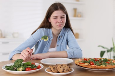 Photo of Sad teen girl holding fork with broccoli at home, selective focus. Eating disorder