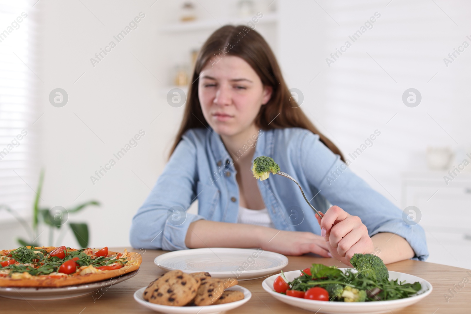 Photo of Sad teen girl holding fork with broccoli at home, selective focus. Eating disorder