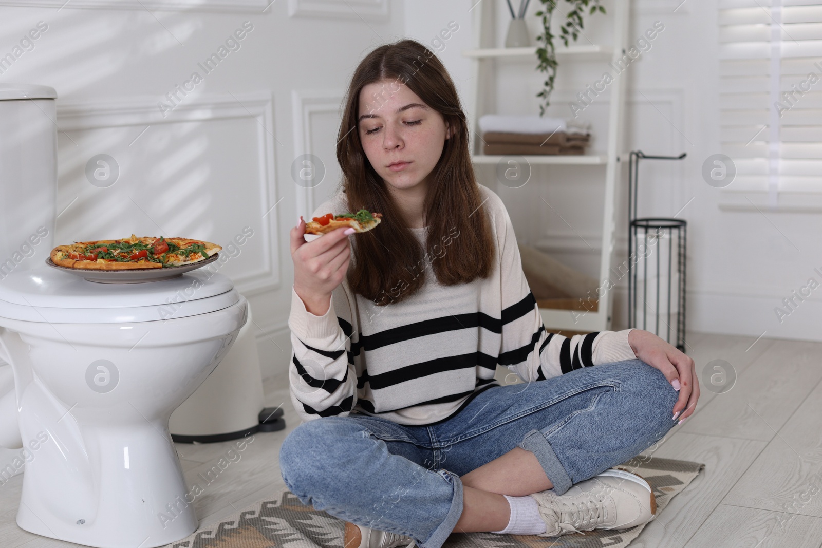 Photo of Teen girl eating pizza near toilet bowl at home. Bulimia