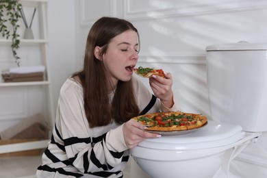 Photo of Teen girl eating pizza near toilet bowl at home. Bulimia